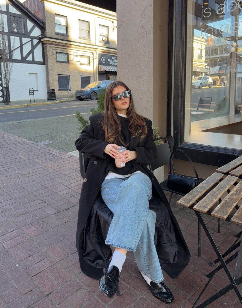 Woman sitting outside a café in a long black wool coat, black sweater, light-wash wide-leg jeans, white socks and black leather loafers