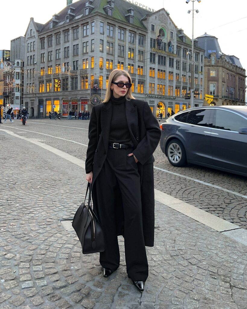 Woman standing in Dam Square Amsterdam wearing a long black coat over a black blazer, black turtleneck, wide-leg black trousers and pointed boots