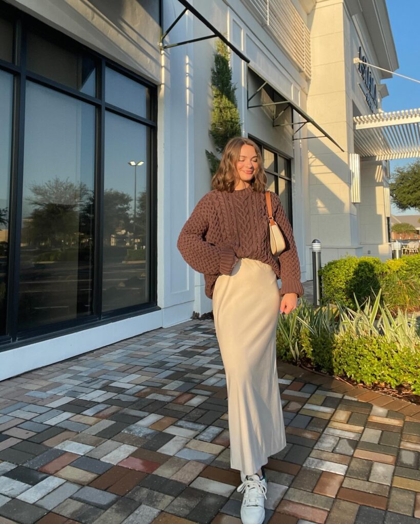Petite woman in a chunky brown cable-knit sweater tucked into a cream satin slip skirt with white sneakers, golden-hour fall light