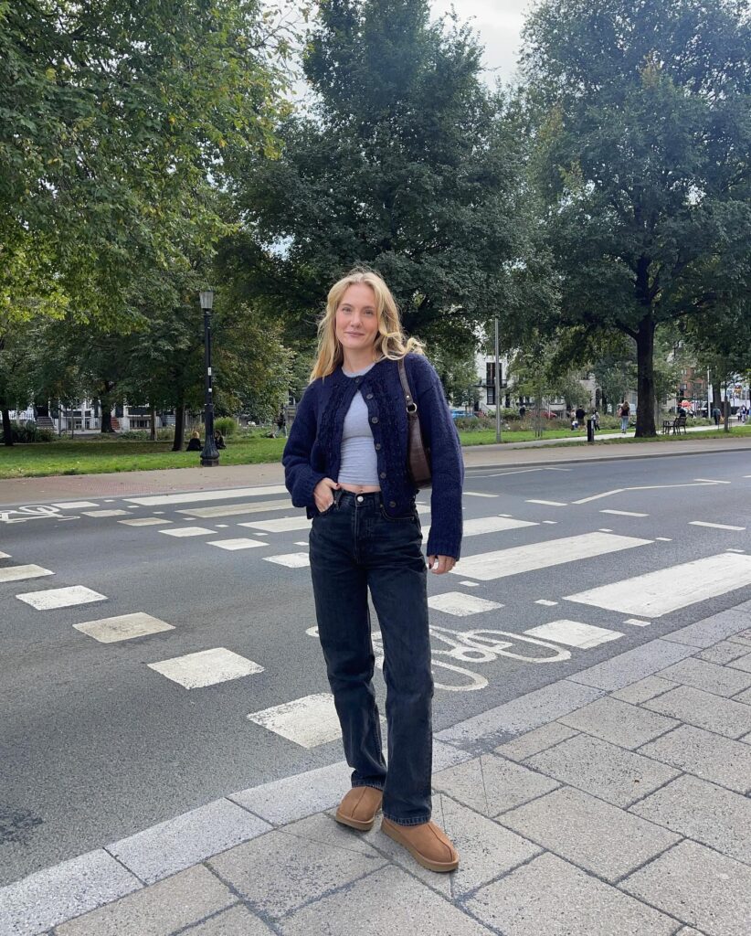 Petite woman in a navy textured cardigan buttoned over a gray tee with black jeans and tan cozy mini boots, crossing a street in fall