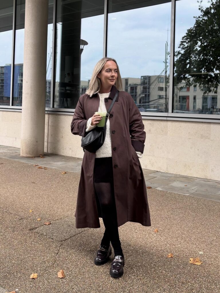 Petite woman in a rich brown trench over a cream knit with a black skirt, dark tights and chunky loafers on a paved walkway