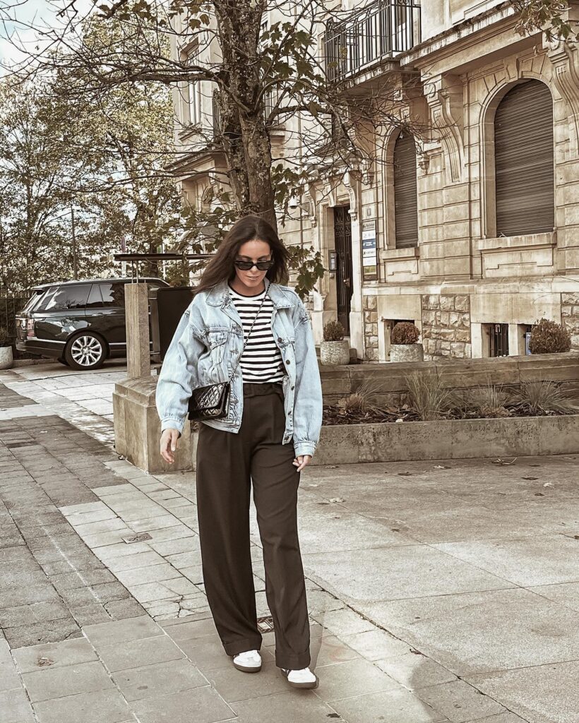 Petite woman in an oversized light-wash denim jacket over a black-and-white striped tee with brown wide-leg trousers and white sneakers