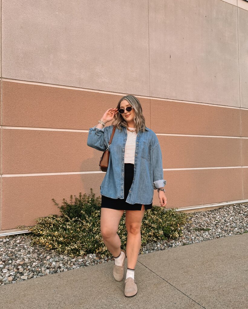 Petite woman in an open chambray shirt over a striped tee with a short black skirt, socks and tan clogs against a striped wall