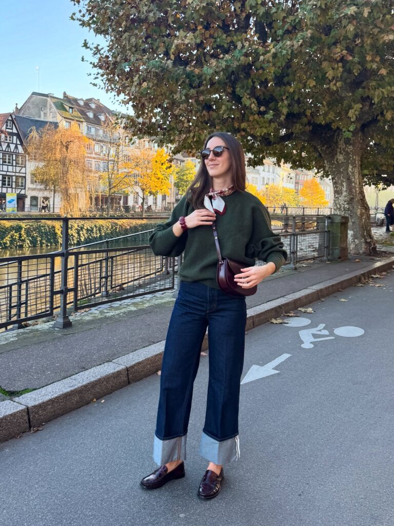 Petite woman in an olive-green sweater with a silky neck scarf, dark cuffed wide-leg jeans and burgundy loafers by an autumn canal