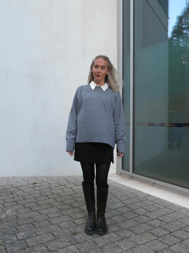 Petite woman in a gray sweater with a crisp white collar peeking out, a short black skirt, dark tights and black knee-high boots against a white wall
