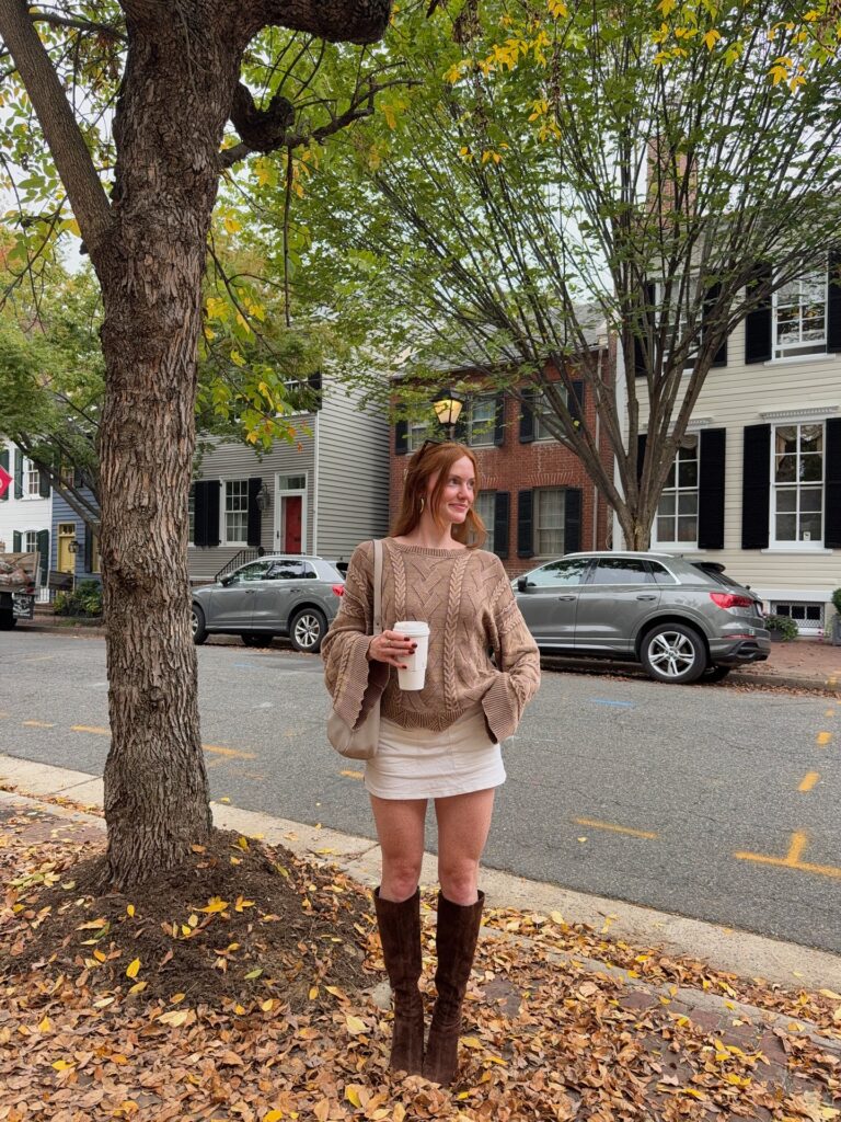 Petite woman in a soft tan cable sweater with bell sleeves, a short white skirt and brown suede knee-high boots on a leafy fall street