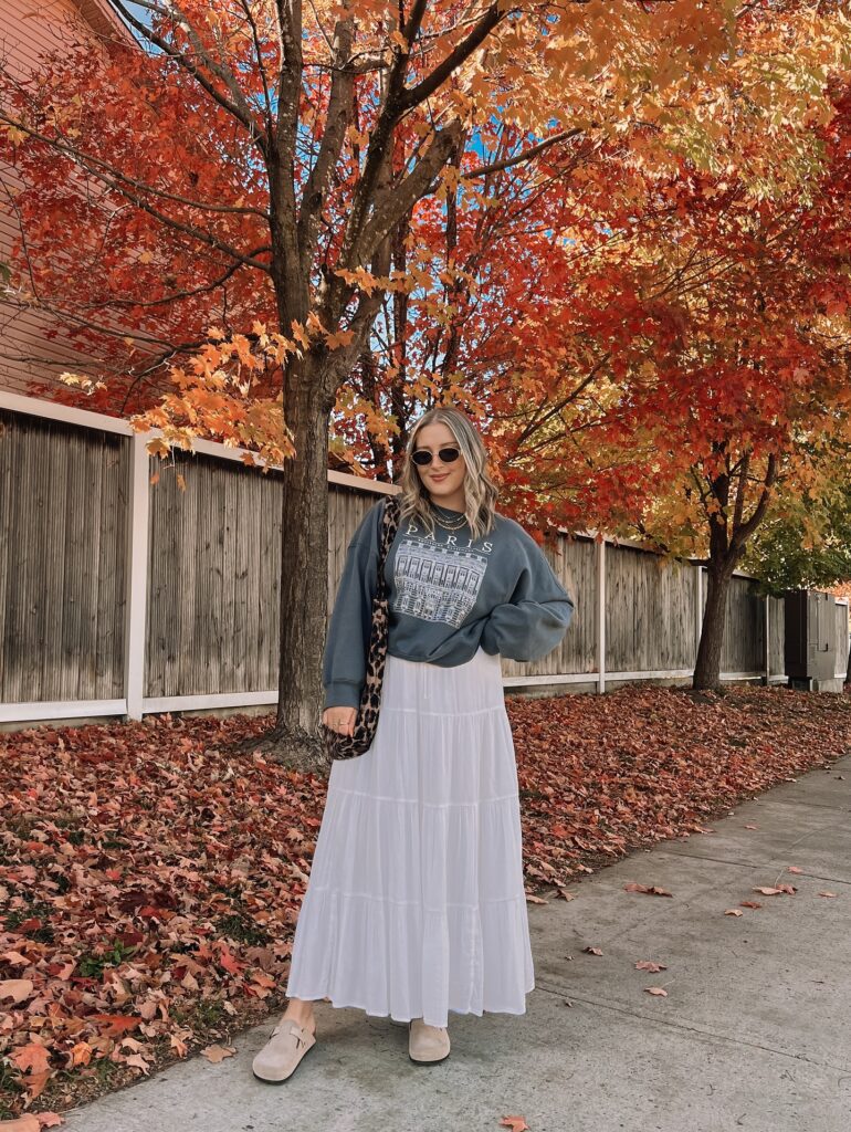 Petite woman in a vintage gray Paris sweatshirt over a white tiered maxi skirt with suede clogs and a leopard-print bag, vivid orange fall foliage behind
