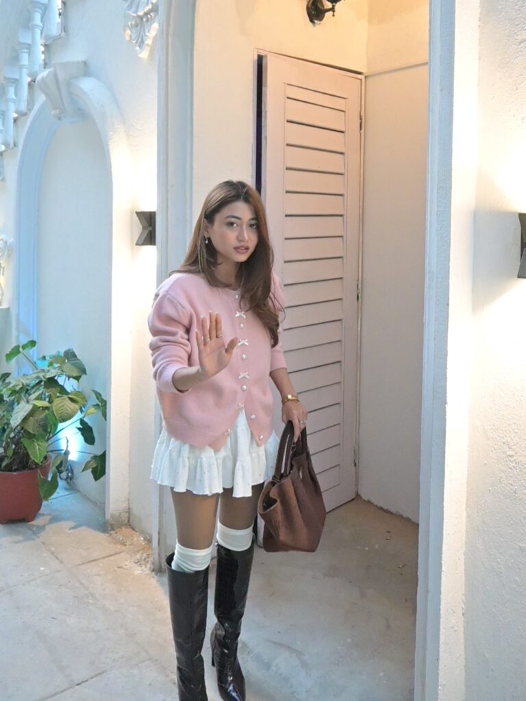 Petite woman in a pink bow-button cardigan, white ruffle mini skirt, white over-the-knee socks and black tall boots in a bright courtyard