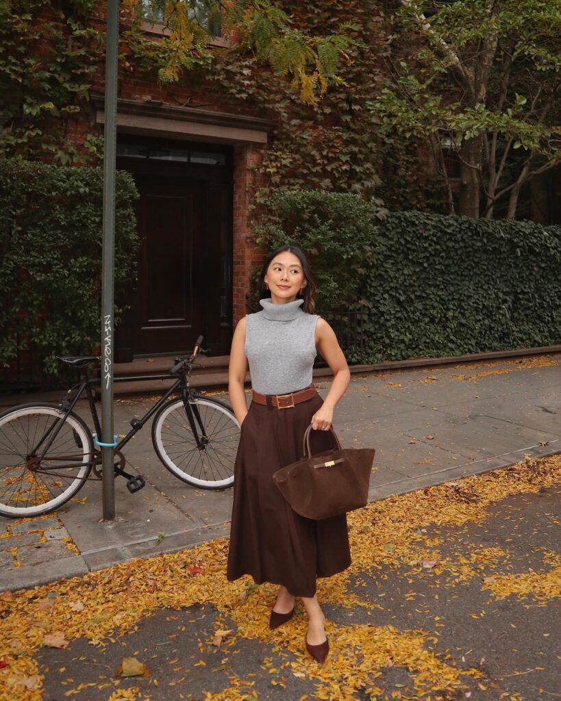 Petite woman in a fitted gray sleeveless turtleneck, brown belt and long brown A-line skirt with pointed flats, autumn leaves underfoot