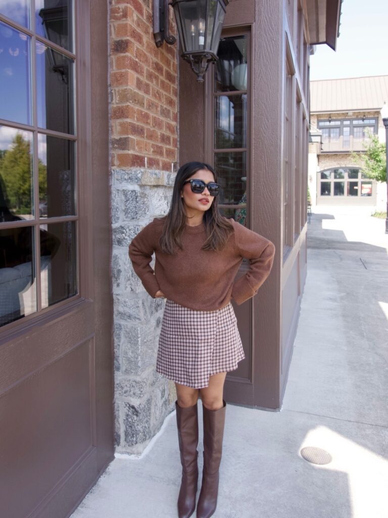 Petite woman in a brown knit sweater, a brown-and-white plaid mini skirt and brown knee-high boots in warm evening light