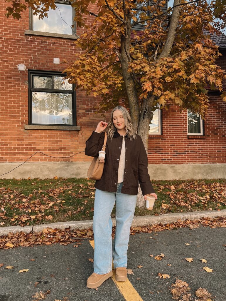 Petite woman in a brown chore jacket over a cream knit with light-wash straight jeans and tan platform clogs, autumn leaves and brick behind her