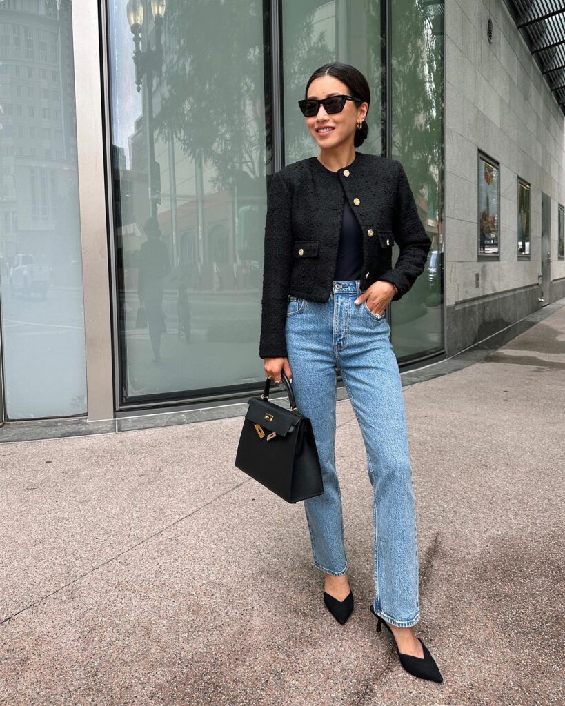 Petite woman in a black tweed jacket with gold buttons over a navy top, straight light-wash jeans and black pointed heels, holding a structured black bag