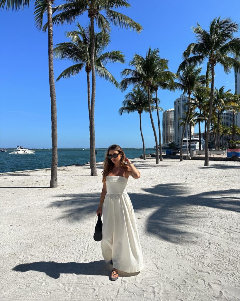 Woman in a white strapless cotton maxi dress holding a black bag, walking on white sand with palm trees and the Miami bay behind her