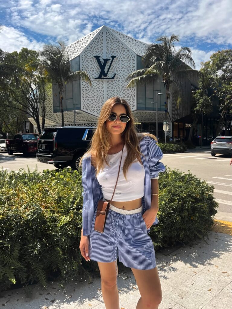 Woman in a blue-and-white striped shirt and matching shorts set worn open over a white crop top with a brown crossbody, palm trees behind her