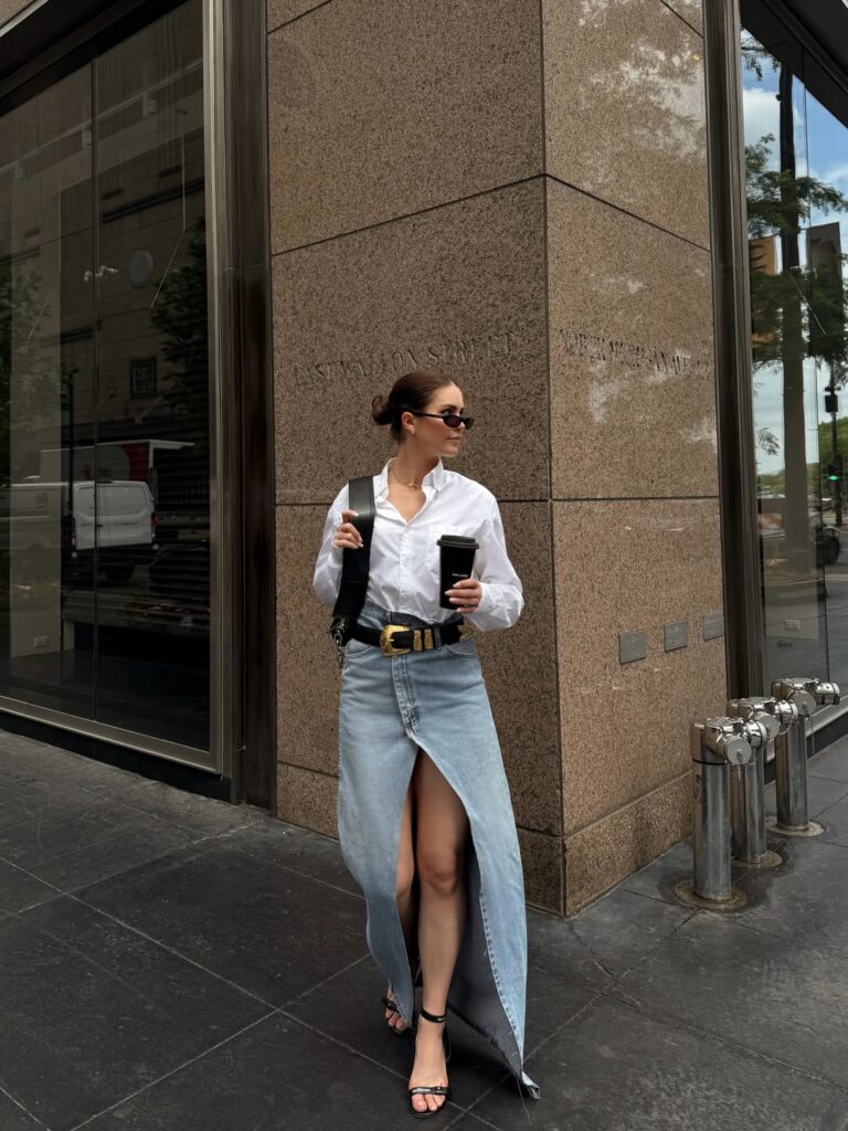 Woman in a crisp white button-down tucked into a light-wash denim maxi skirt with a high slit, black sandals and a takeaway coffee