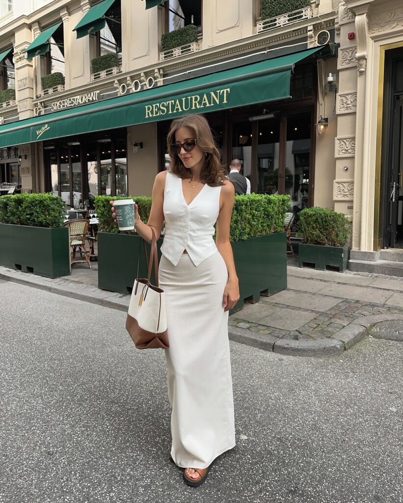 Woman in a white tailored waistcoat and cream column maxi skirt holding a coffee and a slouchy brown leather tote on a city street