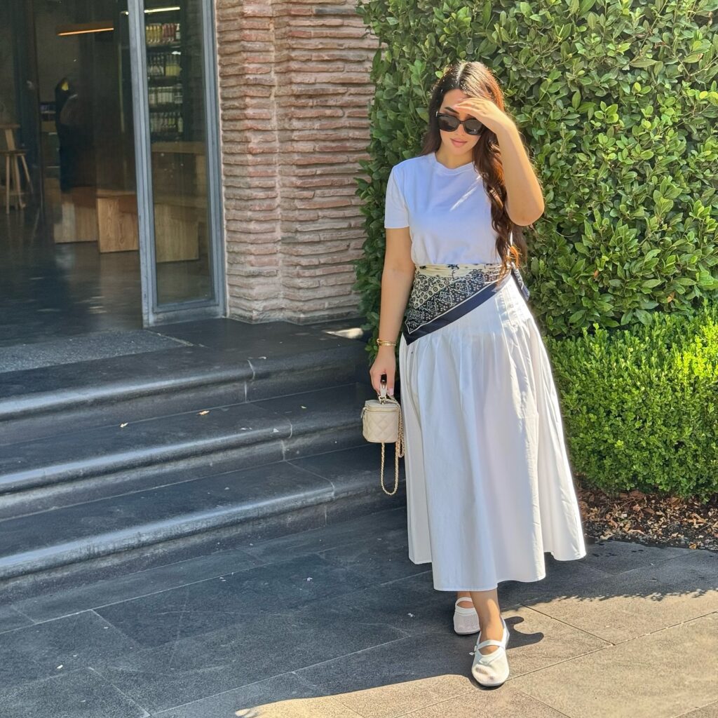 Woman in a plain white tee and voluminous white cotton midi skirt with a printed scarf tied at the waist, ballet flats and a small quilted bag