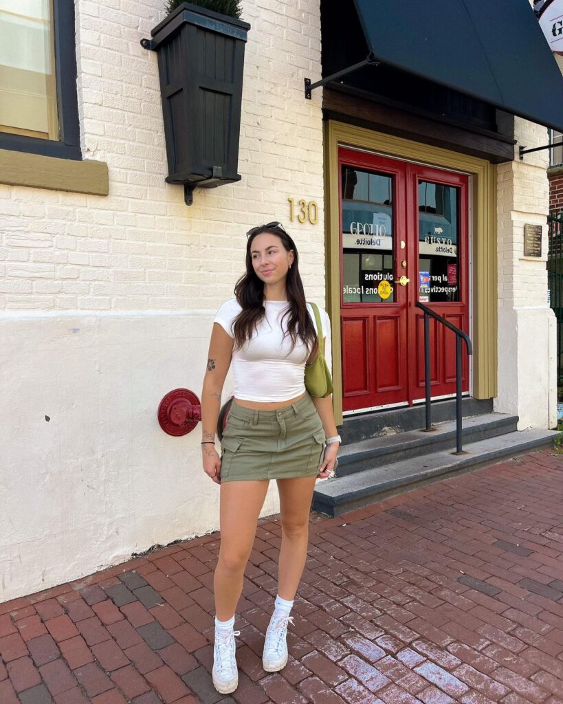 Woman in a white baby tee and olive-green cargo mini skirt with white high-top sneakers and a green shoulder bag in front of a red door