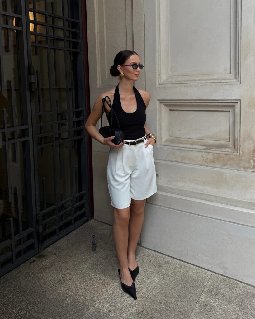Woman leaning against a doorway in a black halter tank and tailored white bermuda shorts with black kitten-heel mules and a small black bag