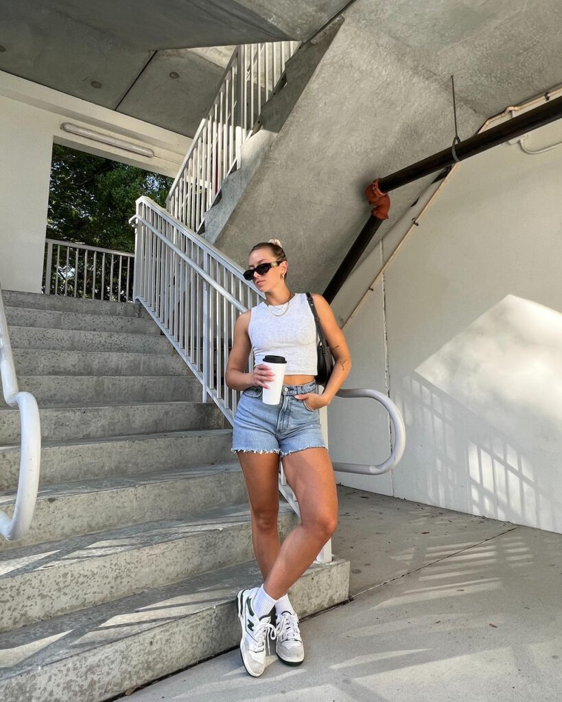 Woman leaning on a stairwell railing in a fitted grey tank and light-wash denim cutoff shorts with white-and-green New Balance sneakers and a black bag