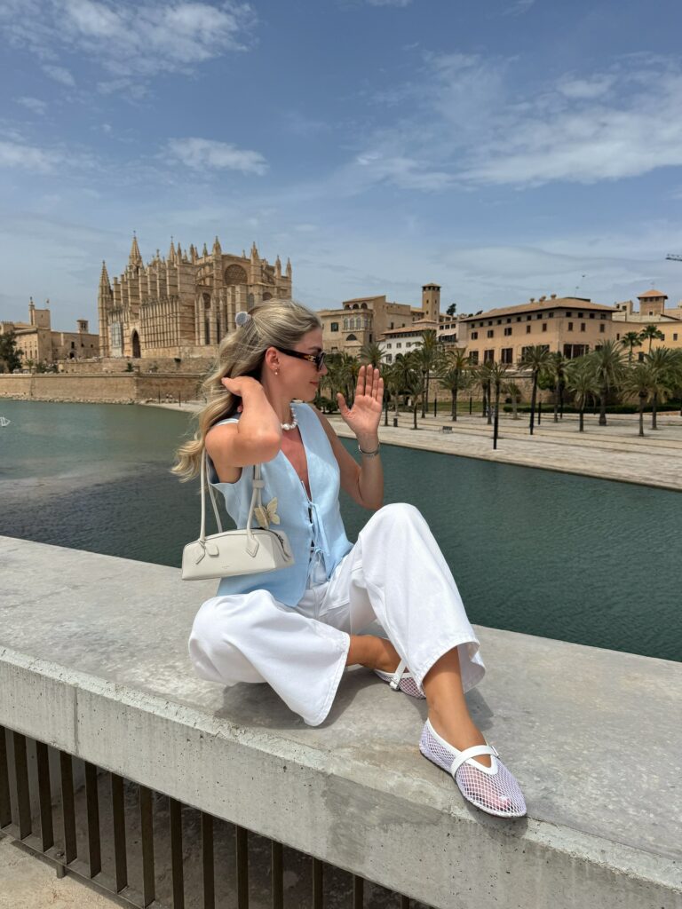 Woman sitting on a ledge in a pale-blue linen waistcoat tied at the front and white wide-leg jeans with a structured white bag and white mesh flats, waterfront behind her