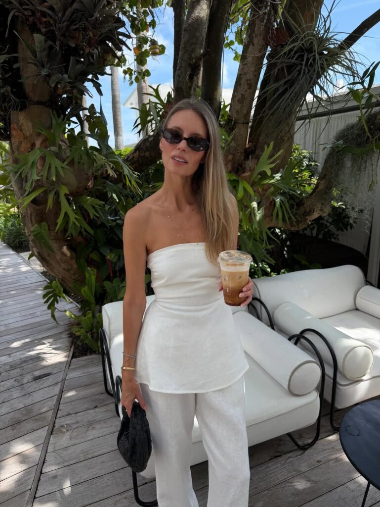 Woman in a white strapless linen top and matching white trousers holding an iced coffee and a black woven bag on a leafy patio