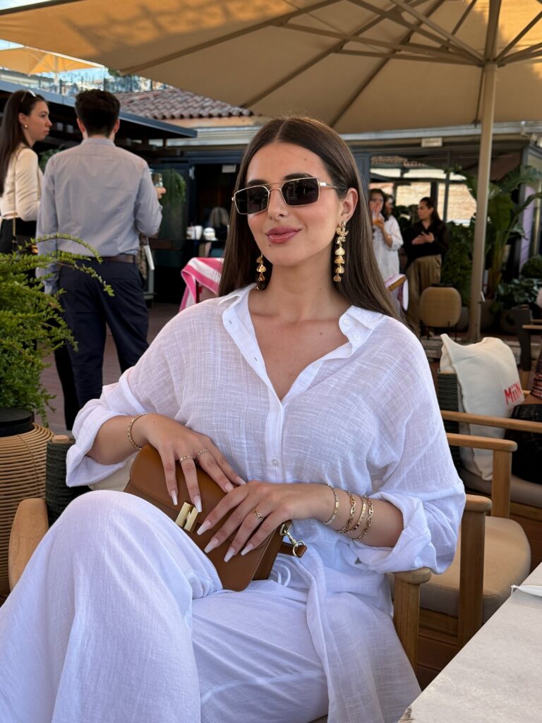 Woman seated at a rooftop bar wearing an oversized sheer white linen shirt and white trousers with a tan leather bag and gold statement earrings
