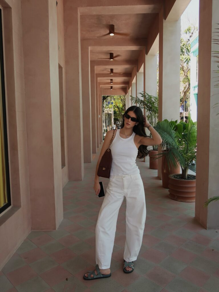 Woman in a white ribbed tank and slouchy white jeans with a brown Chanel flap bag and tan dad sandals in a pink-toned colonnade