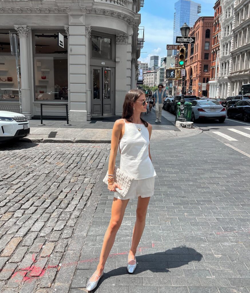 Woman crossing a cobblestone SoHo street in a white square-neck top and white shorts with silver ballet flats and a woven clutch