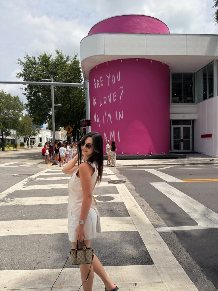 Woman on a crosswalk in a white halter top and tailored white shorts with a small Gucci bag in front of a pink Miami mural