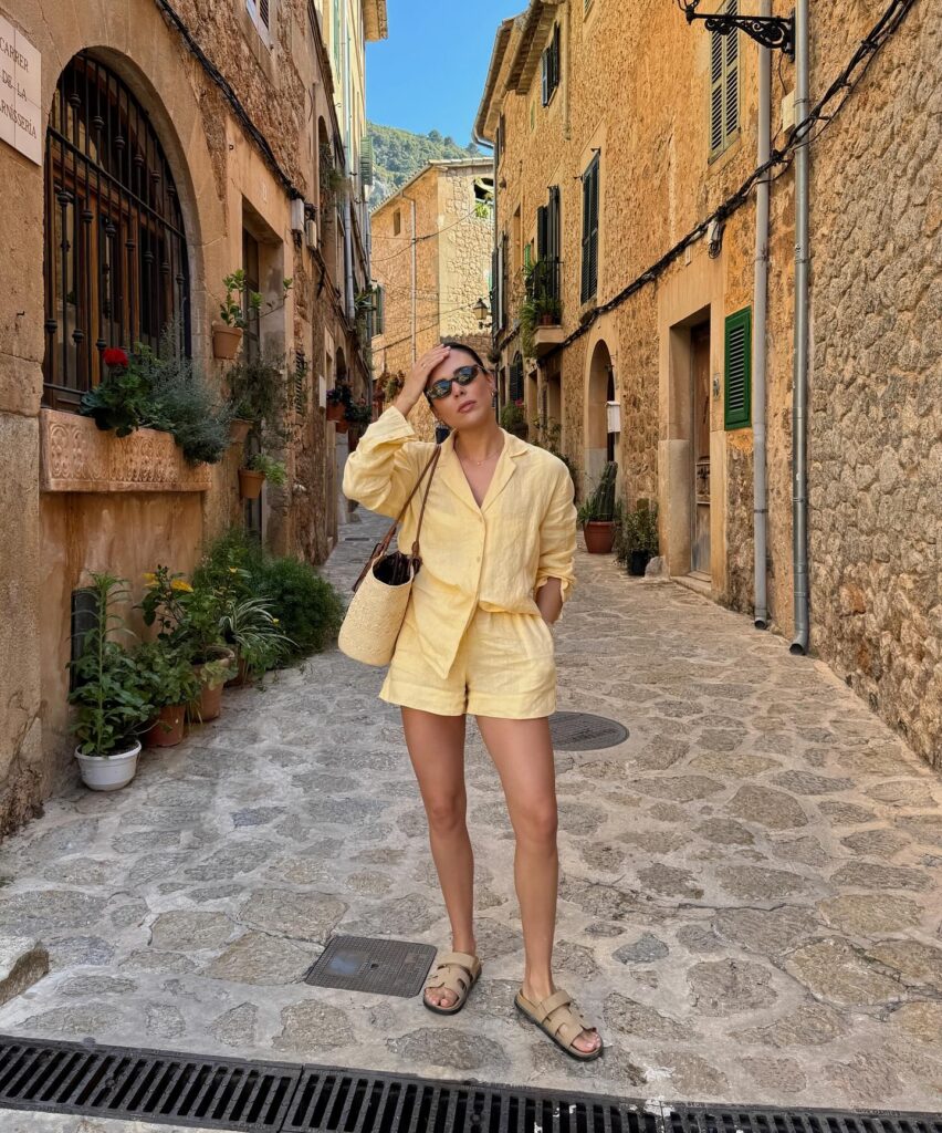 Woman in a soft yellow linen shirt and matching shorts set with tan sandals and a straw bag standing in a sunny stone alley