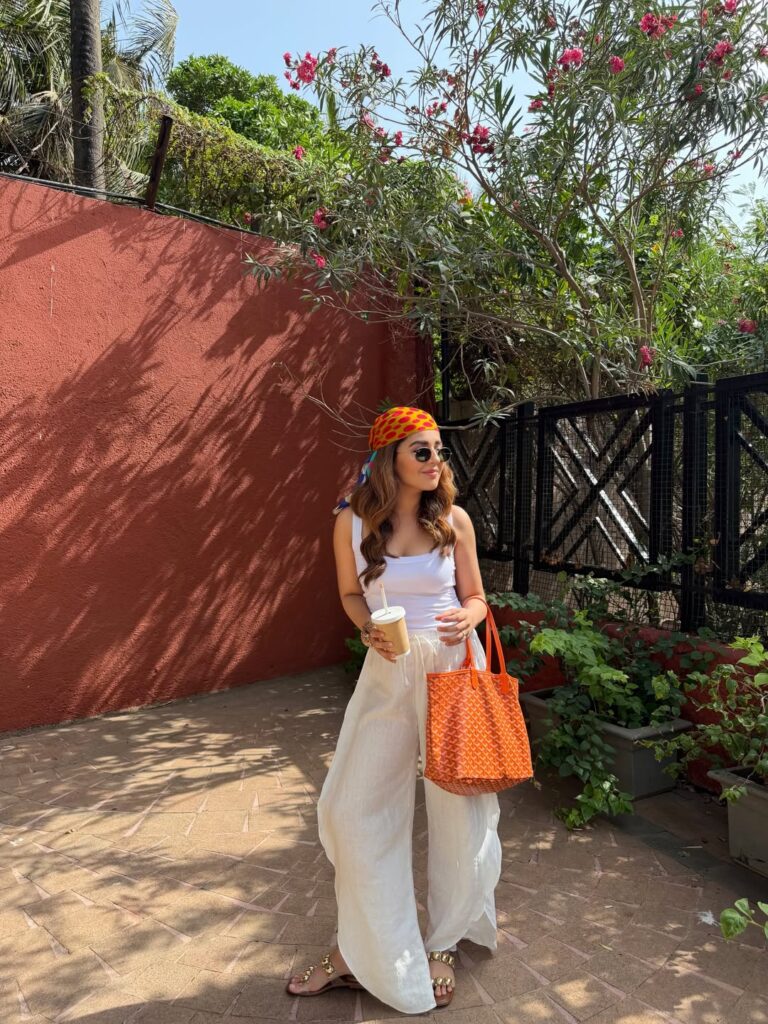 Woman in a white tank and wide white linen trousers with a printed headscarf, orange tote bag and iced coffee against a terracotta wall